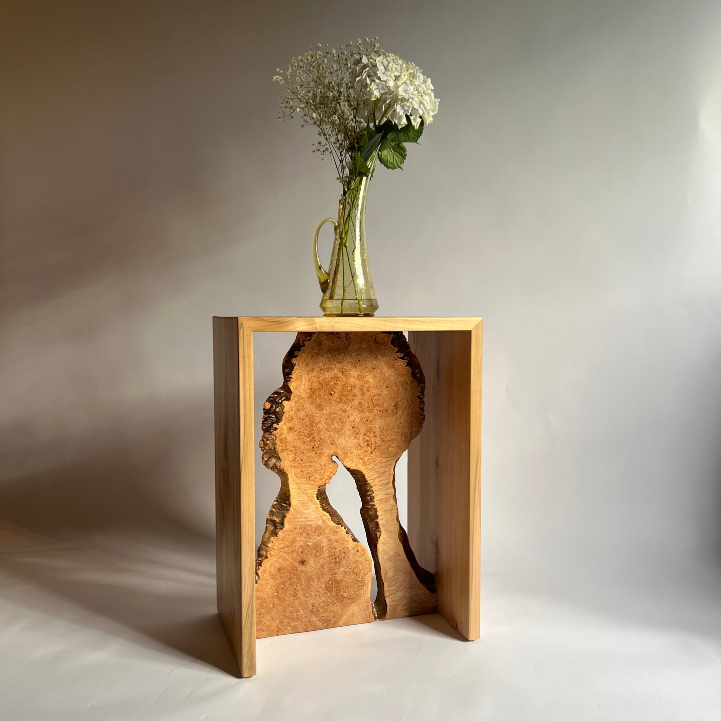 A light natural wood side table with a sculptural burl wood inlay, styled with a glass vase of white flowers on top, set against a neutral gray studio background.
